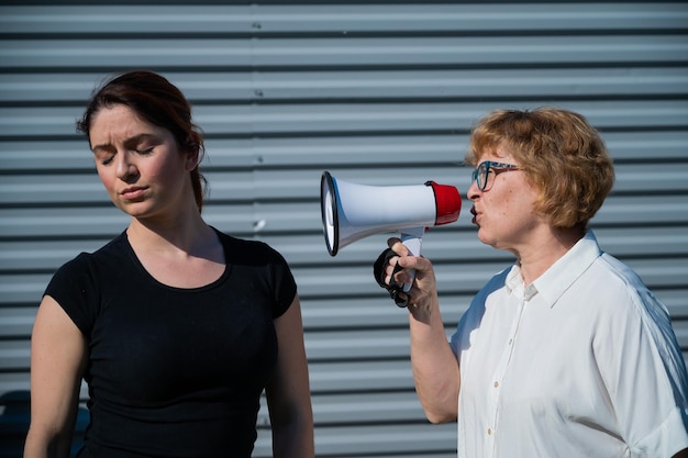 The conflict of generations An old woman yells into a megaphone at her middleaged daughter against gray background Quarrel between an elderly mother and daughter