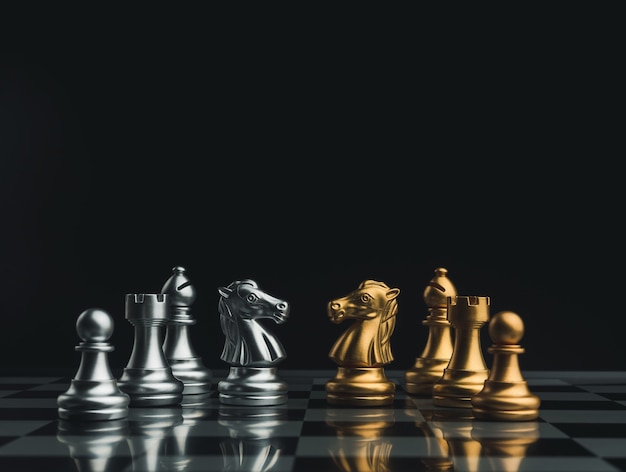 Confrontation of a golden and a silver team of chess pieces, knight, bishop, rook, and pawn on a chessboard on dark background. Leadership, partnership, team, competitor and business strategy concept.