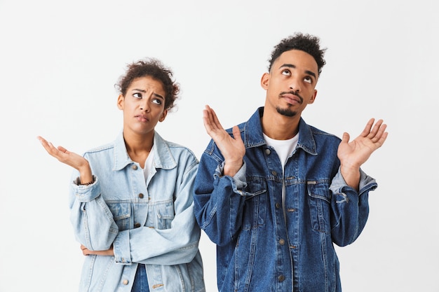 Confused african couple in denim shirts standing together and looking up over grey wall