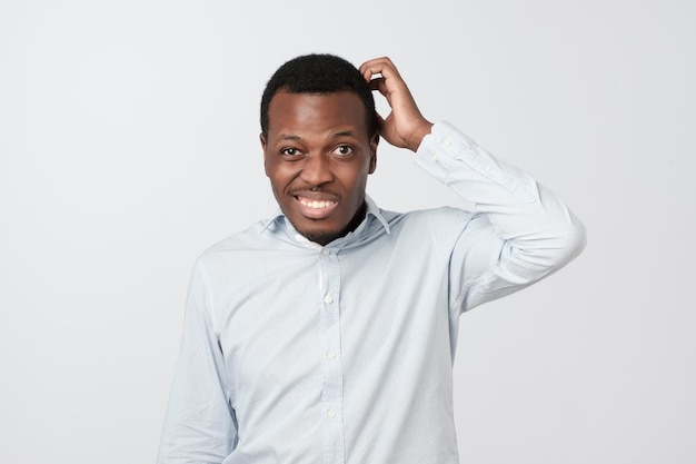 Photo confused african man scratching head while looking at camera and standing over gray background