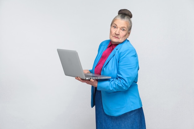 Confused aged woman standing and holding laptop thinking and looking at camera. Grandmother in light blue suit with collected gray hair bun hairstyle. indoor, Studio shot, isolated on gray background