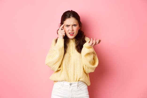 Confused and annoyed girl pointing at head with index finger, scolding someone for stupid or crazy behaviour, being disappointed, standing against pink wall.