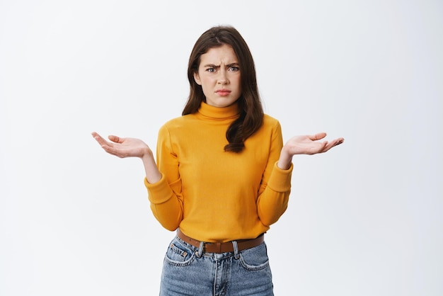 Confused and annoyed young woman frowning raising hands sideways and shrugging clueless cant understand what happened dont know anything standing against white background