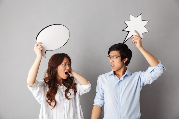 Confused asian couple standing isolated, holding empty speech bubble, having an argument