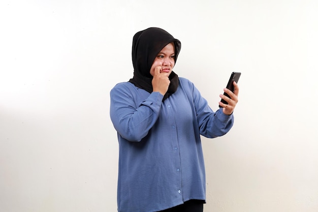 Сбитая с толку азиатская мусульманка, стоящая с мобильным телефоном, изолирована на белом