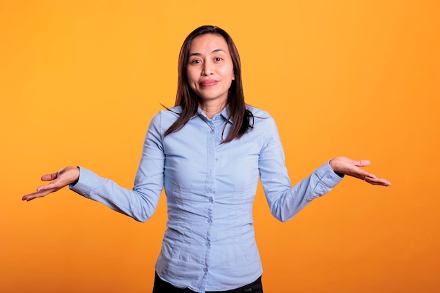 Confused asian woman being hesitant and unconfident while trying to answering question. doubtful young adult shrugging shoulders having clueless expression in studio over yellow background