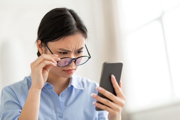 Confused asian woman holding smartphone removing eyeglasses