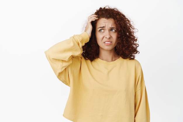 Confused awkward girl with curly hair, scratching head and looking aside clueless, dont understand, has no idea, puzzled, stands indecisive against white background.