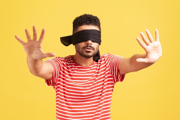 Confused bearded man in red striped tshirt with blindfold on eyes moving in darkness with outstretched arms training developing senses Indoor studio shot isolated on yellow background