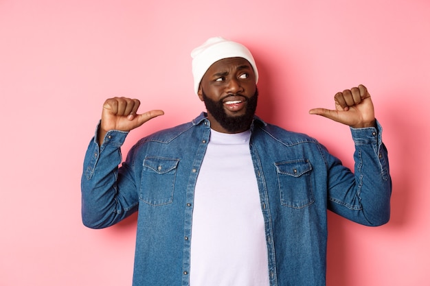 Confused Black man staring right and pointing at himself unsure, having doubts, standing over pink background