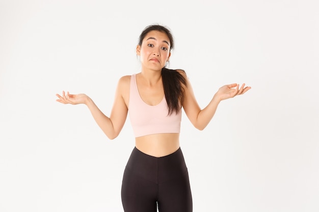 Confused and clueless asian fitness girl, female athlete in sportswear dont know, shrugging and grimacing puzzled, standing unaware white background.