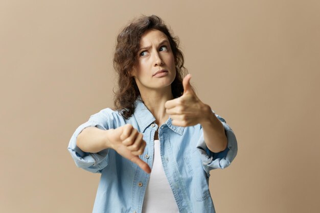 Confused curly beautiful female in jeans casual shirt can not make decision show thumbs up down looks up posing isolated on over beige pastel background People Lifestyle emotions concept Copy space