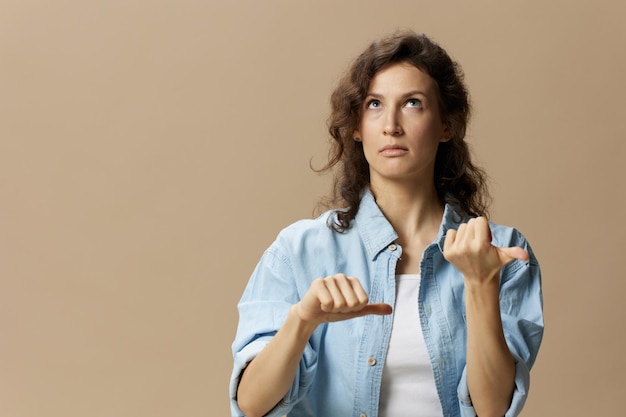 Confused curly beautiful female in jeans casual shirt can not make decision show thumbs up down looks up posing isolated on over beige pastel background People Lifestyle emotions concept Copy space