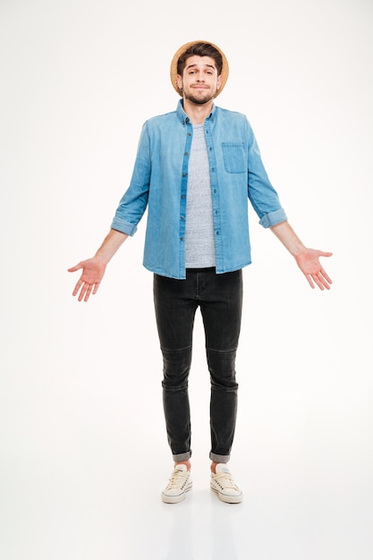 Фото Смущенный милый молодой человек в джинсовой рубашке стоит и пожимает плечами на белом фоне