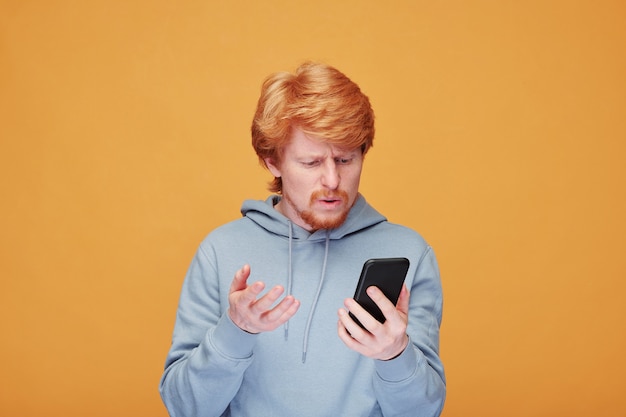 Confused or dissatisfied young man in hoodie talking to someone while looking at screen of smartphone over yellow wall