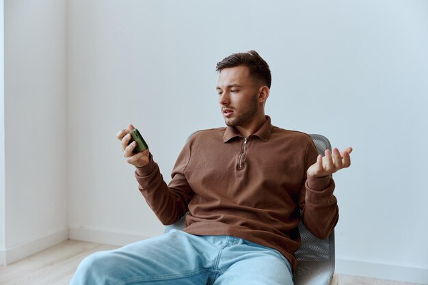 Confused excited thoughtful young tanned man don39t know what\
answer raise palm up with question hold phone sitting on chair at\
home near white wall distance communication concept copy space