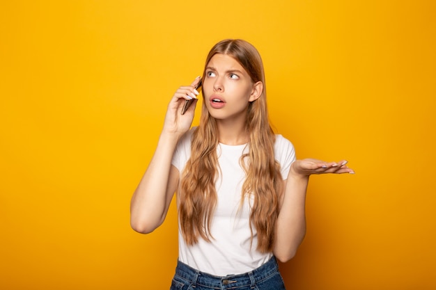 Confused girl showing shrug gesture while talking on smartphone isolated on yellow