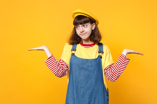 Confused girl teenager in french beret, denim sundress spreading, pointing hands, looking aside isolated on yellow background in studio. People sincere emotions, lifestyle concept. Mock up copy space.