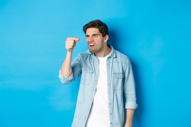 Confused guy holding something tiny in fingers and looking at it puzzled, standing over blue wall