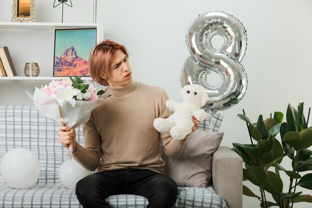 Confused handsome guy on happy women day holding bouquet and looking at teddy bear in his hand sitting on sofa in living room