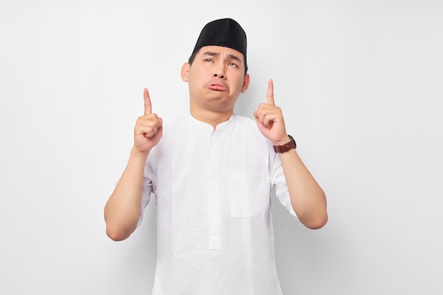 Confused handsome young Asian Muslim man wearing Arabic costume pointing finger at free space place for an advertisement isolated on white background People religious Islamic lifestyle concept