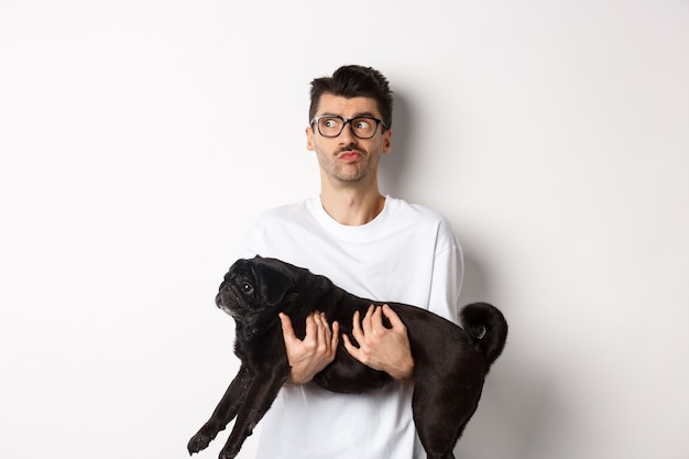 Confused hipster guy holding black cute pug and looking left, feeling puzzled or clueless. Dog owner pondering, carry dog in arms, white.