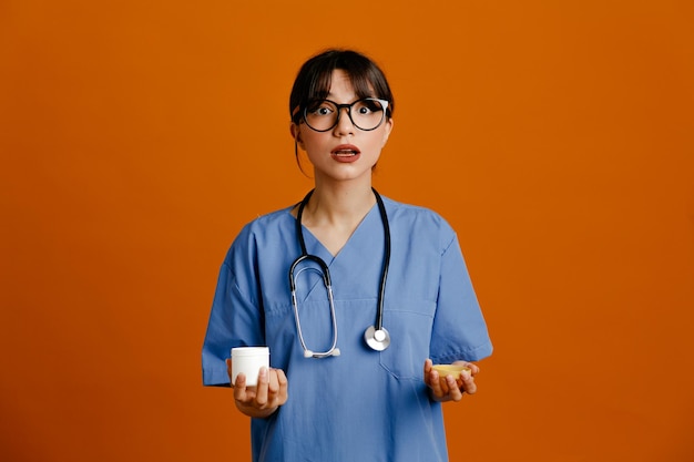 Latta confusa della holding della pillola giovane dottoressa che indossa uno stetoscopio fitth uniforme isolato su sfondo arancione