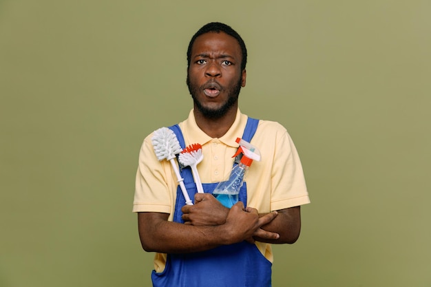 Confused holding cleaning tools young africanamerican cleaner male in uniform with gloves isolated on green background