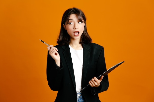 Confused holding clipboard and pen young beautiful female wearing black jacket isolated on orange background