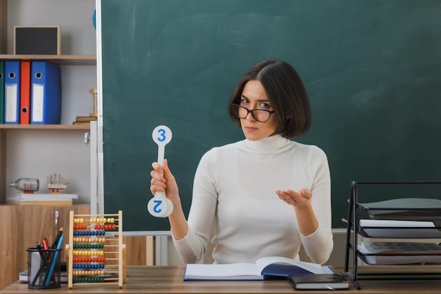 教室で学校の道具をつけて机に座っているナンバーファンを持って眼鏡をかけている若い女性教師がカメラに手をかざすのに混乱しました