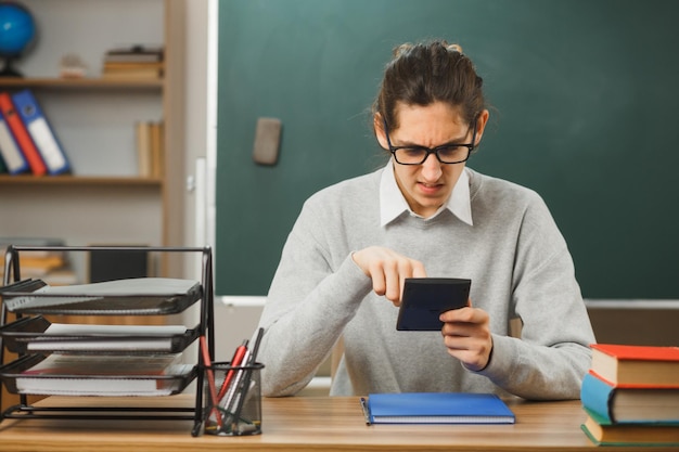 confused holding and looking at calculator young male teacher sitting at desk with school tools on in classroom