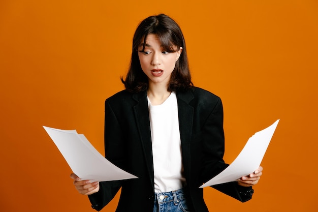 Confused holding papers young beautiful female wearing black jacket isolated on orange background