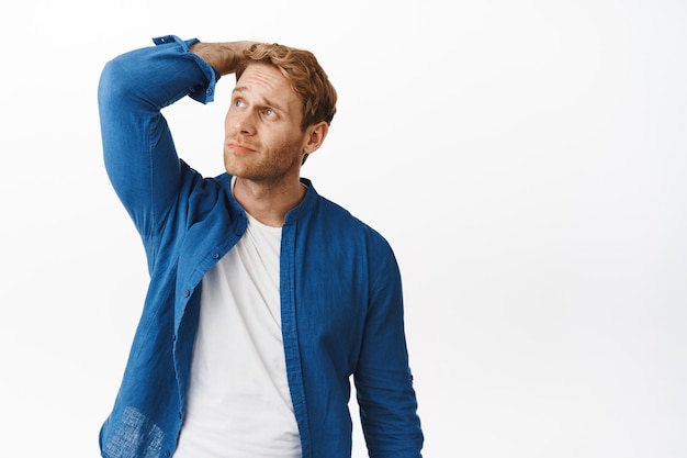 Confused and indecisive redhead guy scratch head, look puzzled at upper left corner, feeling awkward or unsure, standing questioned against white background