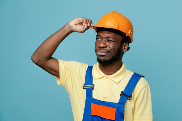 Confused looking at side young african american builder in uniform isolated on blue background