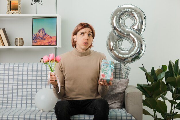 Confused looking up handsome guy on happy women day holding flowers with present sitting on sofa in living room