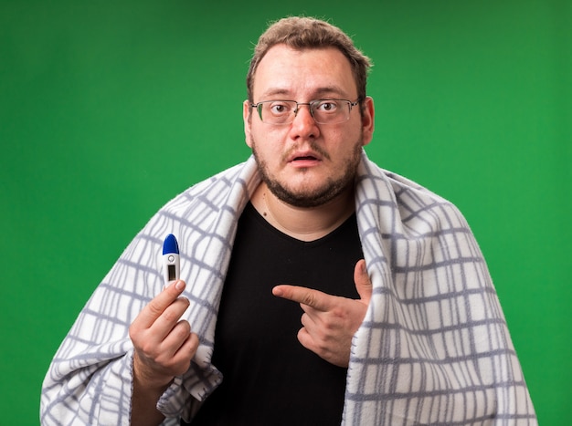 Confused middle-aged ill male wrapped in plaid holding and points at thermometer isolated on green wall