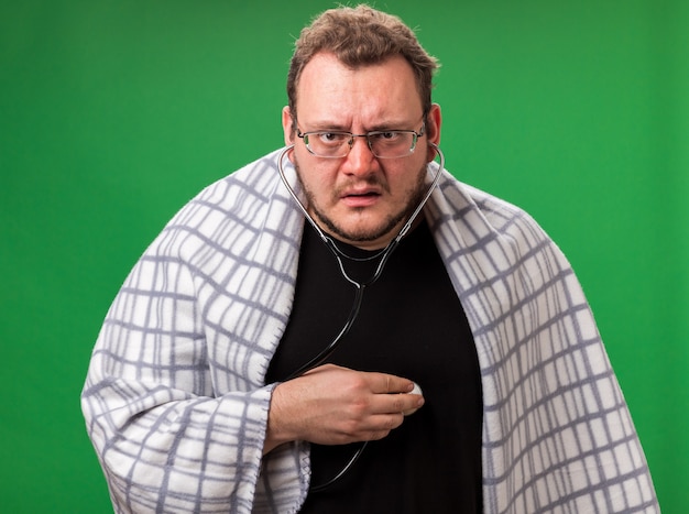 Confused middle-aged ill male wrapped in plaid listening to his own heartbeat isolated on green wall