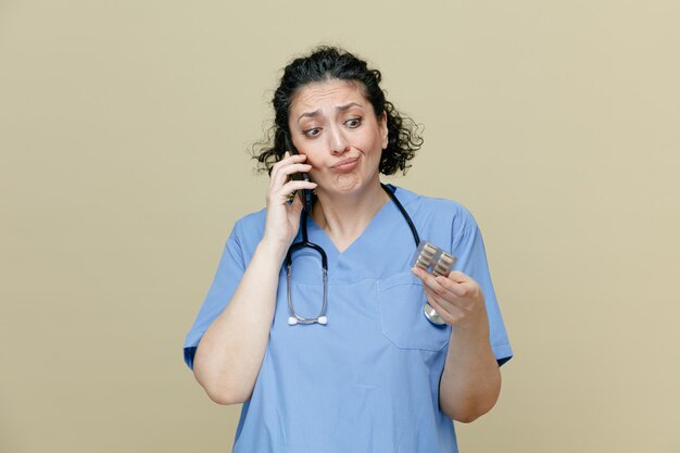 Medico femminile di mezza età confuso che indossa uniforme e stetoscopio intorno al collo che tiene un pacchetto di pillole guardandolo mentre parla al telefono isolato su sfondo verde oliva