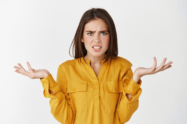 Confused and nervous woman asking what, spread hands sideways and staring at front puzzled, cant understand whats happening, standing over white wall