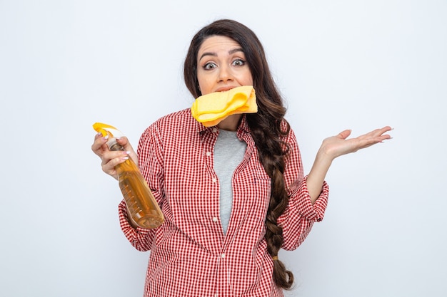 Confused pretty caucasian cleaner woman keeping cleaning cloths with her teeth and holding spray cleaner 