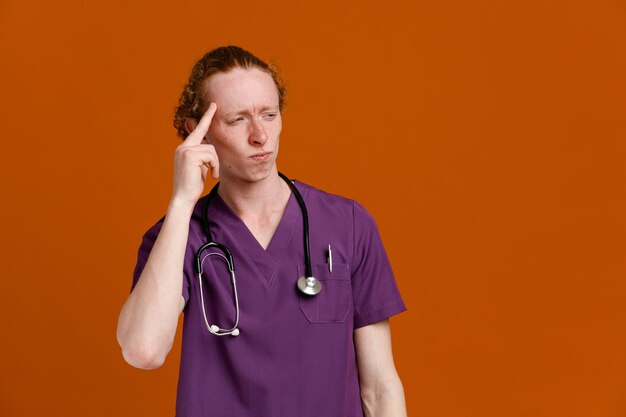 Confused putting finger on head young male doctor wearing uniform with stethoscope isolated on orange background