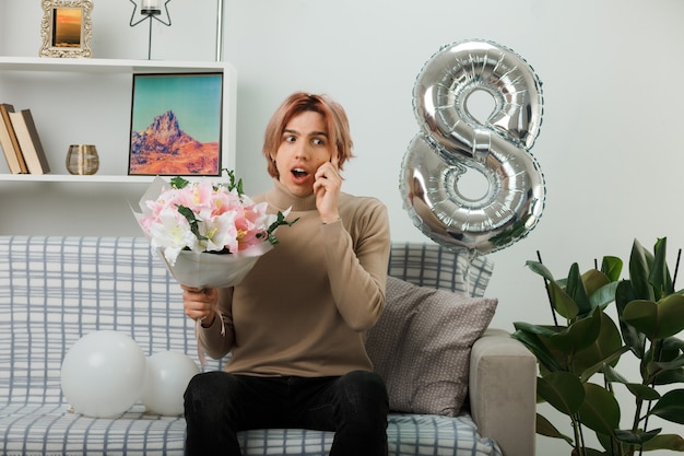 Confused putting hand on cheek handsome guy on happy women day holding bouquet sitting on sofa in living room