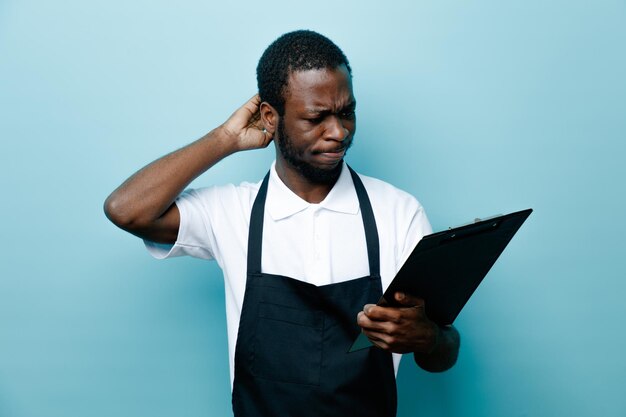 Confused putting hand on head holding clipboard young african american barber in uniform isolated on blue background