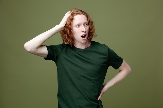 Confused putting hands on head and hips young handsome guy wearing green t shirt isolated on green background