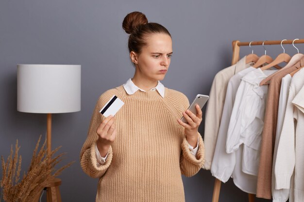 Confused puzzled woman in beige sweater stands against gray wall with clothes hang in wardrobe on rack holding cell phone and credit card has not enough money on her bill can't to pay for purchases