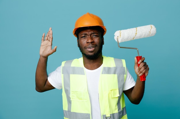 Mano alzata confusa giovane costruttore afroamericano in uniforme che tiene una spazzola a rullo isolata su sfondo blu