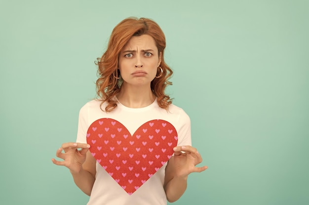 Photo confused redhead girl hold red heart on blue background