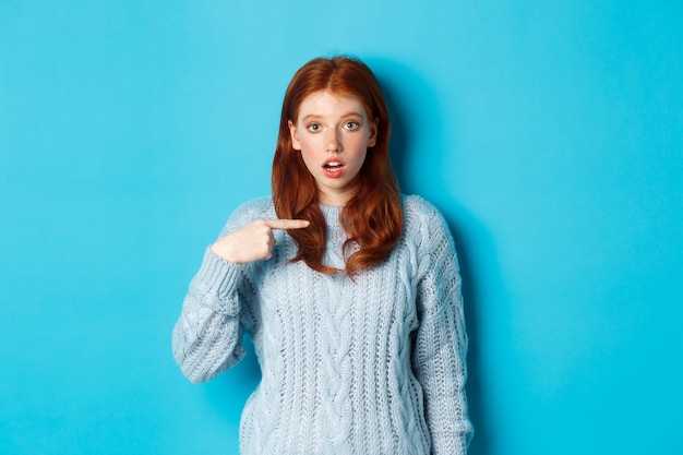 Смущенная рыжая девушка, указывающая на себя, будучи выбранной, стоит в свитере на синем фоне