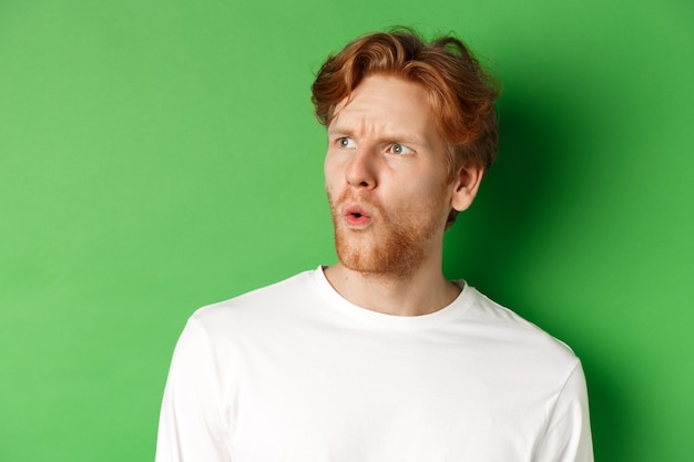 Confused redhead man staring left and looking worried, standing against green background.