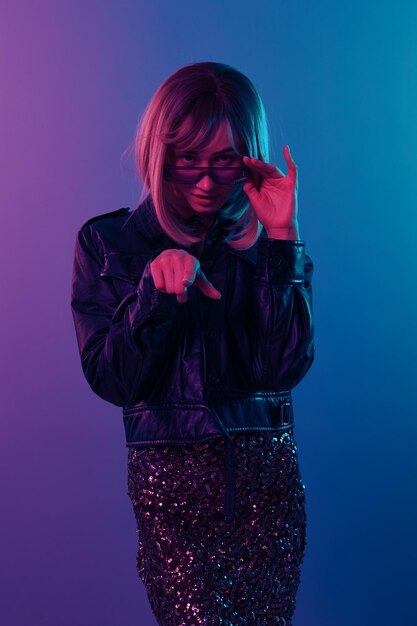 Confused sexy beautiful blonde woman in leather jacket sparkly\
dress trendy sunglasses point finger at camera posing isolated in\
blue pink color light studio background neon party cyberpunk\
concept